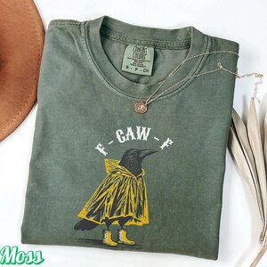 May include: A moss green t-shirt with a graphic of a crow wearing a yellow raincoat and boots. The text "F-GAW-F" is above the crow. A gold necklace with a pendant is draped across the shirt.