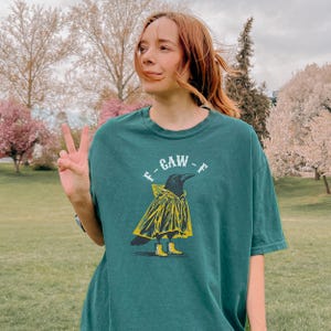 May include: Teal green t-shirt featuring a cartoon crow wearing a yellow raincoat and boots. The text "-GAW-" is above the crow. The shirt is worn by a person outdoors in a park setting.