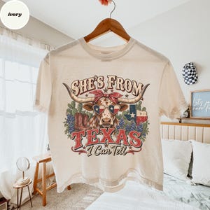 May include: An ivory t-shirt featuring a graphic of a longhorn cow wearing a red bandana and heart-shaped sunglasses with a Texas flag pattern. The design includes cowboy boots, cacti, and the text "SHE'S FROM TEXAS I Can Tell".