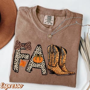 May include: Brown t-shirt with the word "FALL" in a leopard print design, a pumpkin, a bow, and cowboy boots. The shirt also has the word "Espresso" printed on it. A silver necklace with a pendant is on the shirt.