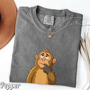 May include: A gray Comfort Colors t-shirt featuring a cartoon monkey with a thoughtful expression. The monkey is brown and tan with a gray hand on its chin. A silver necklace with a small pendant is on the shirt. The word "Pepper" is at the bottom left.