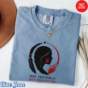 May include: A light blue short-sleeve t-shirt featuring a black silhouette of a person's profile with a red handprint over the mouth and a red handprint below. A black feather and red circular arc surround the graphic with the text NOT INVISIBLE. NOT FORGOTTEN.