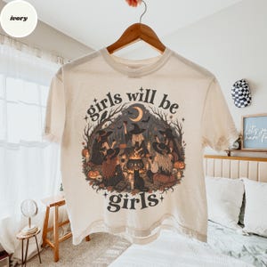 May include: An ivory short-sleeve t-shirt featuring a graphic illustration of five witches gathered around a cauldron in a forest. The text "girls will be girls" is printed in a dark font above and below the central circular design.