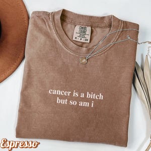 May include: Espresso-colored Comfort Colors t-shirt with the text "cancer is a bitch but so am i" in white. A silver necklace with a small pendant rests on the shirt. The shirt is folded and displayed on a white surface.