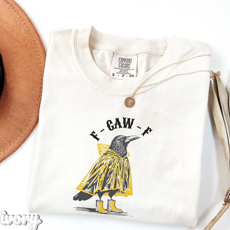 May include: Ivory-colored t-shirt with a graphic of a crow wearing a yellow raincoat and boots, with the text "F-GAW-F". A gold necklace and a brown hat are also visible.