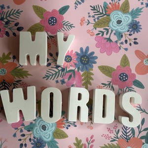 Op de afbeelding: Witte blokletters die "MY WORDS" spellen op een roze bloemenachtergrond. De bloemen zijn in tinten roze, blauw en oranje met groene bladeren. De letters zijn gelijkmatig verdeeld over de afbeelding.