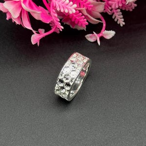 Peut inclure: Une bague en argent avec un motif en nid d'abeille. La bague est posée sur un fond noir avec des fleurs roses et blanches en arrière-plan. Le motif de la bague est un motif géométrique répétitif.