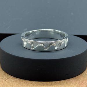 Peut inclure: Une bague en argent avec un motif de vagues. La bague est de forme circulaire et présente un motif répétitif de vagues stylisées sur sa circonférence. Elle est présentée sur un support cylindrique noir, sur fond neutre.