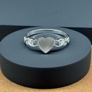 Peut inclure: Une bague argentée avec un sertissage en forme de cœur. La bande présente un motif de nœud celtique complexe. La bague est présentée sur un support circulaire gris foncé, avec un fond neutre.