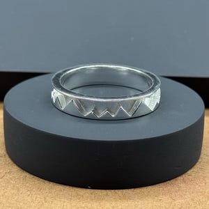 Peut inclure: Une bague en argent avec un motif de chaîne de montagnes. La bague présente une série de pics triangulaires autour de la bande. La bague est exposée sur un support circulaire noir.