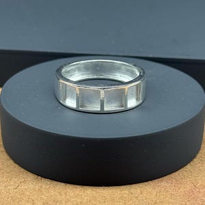 Peut inclure: Une bague argentée avec un motif rectangulaire. La bague est en métal brillant et comporte une série de découpes rectangulaires autour de l'anneau. La bague est présentée sur un socle circulaire noir sur fond neutre.
