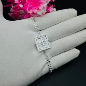 Puede incluir: Una pulsera de plata con una pieza central cuadrada con cuatro cuadrados más pequeños. La pulsera tiene un diseño de cadena delicado. La joya se exhibe sobre una superficie blanca con flores rosas en el fondo.
