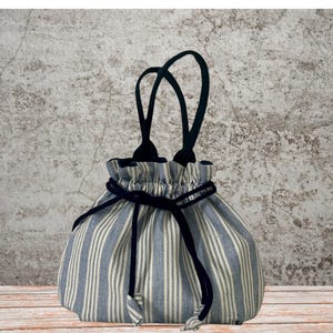 Peut inclure: Un sac à cordon bleu et blanc rayé avec des poignées noires et une fermeture à cordon noire. Le sac a une forme arrondie et est fait de tissu. Les poignées sont attachées au sommet du sac.