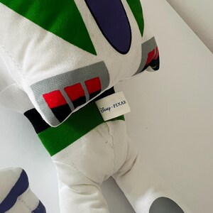 May include: A plush toy in the likeness of Buzz Lightyear from Disney Pixar's Toy Story. The toy features a white body with green, purple, gray, and red accents. The Disney Pixar tag is visible.