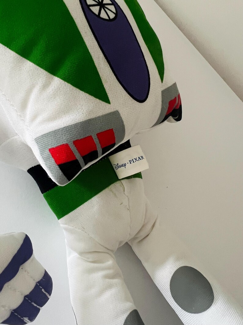 May include: A plush toy in the likeness of Buzz Lightyear from Disney Pixar's Toy Story. The toy features a white body with green, purple, gray, and red accents. The Disney Pixar tag is visible.