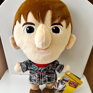 May include: A plush doll of a character with brown hair, blue eyes, and a tan face. The doll wears a camouflage jacket over a red shirt and brown shoes. A yellow tag with the text "Only Fools and Horses" is attached.