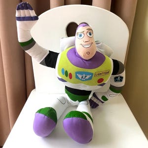 May include: A plush Buzz Lightyear doll, featuring a purple and white head, a green and yellow chest, and white limbs with green and purple accents. The doll is sitting on a white chair and has the word "LIGHTYEAR" on its chest.