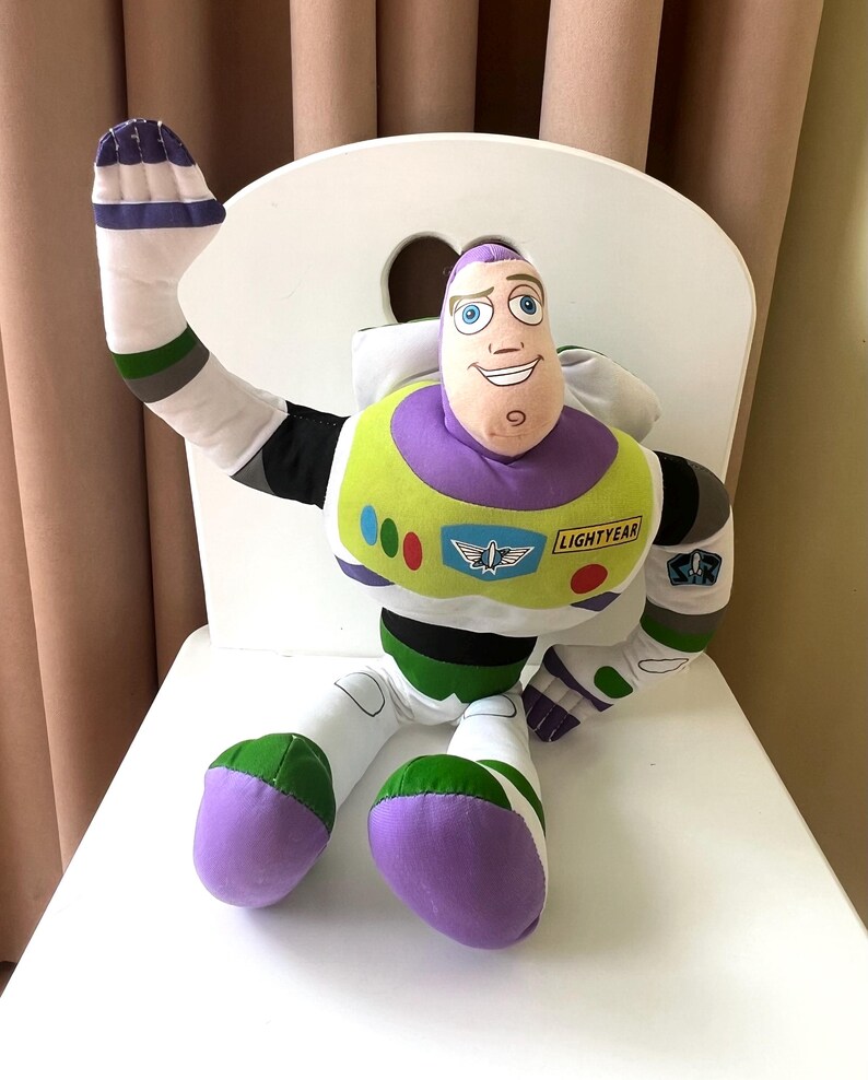 May include: A plush Buzz Lightyear doll, featuring a purple and white head, a green and yellow chest, and white limbs with green and purple accents. The doll is sitting on a white chair and has the word "LIGHTYEAR" on its chest.