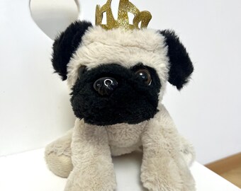 May include: A plush pug dog toy with a black muzzle and ears, wearing a gold glitter crown. The toy is beige and black, with large, expressive eyes. It is sitting on a white surface.