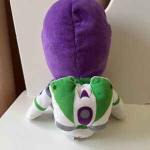 Pode incluir: Um brinquedo de pel&uacute;cia do Buzz Lightyear, com cabe&ccedil;a roxa, fato espacial branco e verde e detalhes cinzentos. O brinquedo est&aacute; sobre uma superf&iacute;cie branca.