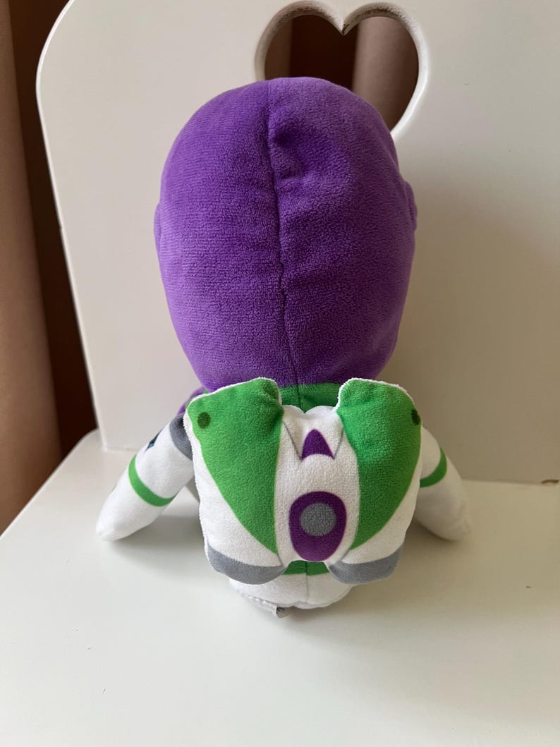 Pode incluir: Um brinquedo de pel&uacute;cia do Buzz Lightyear, com cabe&ccedil;a roxa, fato espacial branco e verde e detalhes cinzentos. O brinquedo est&aacute; sobre uma superf&iacute;cie branca.