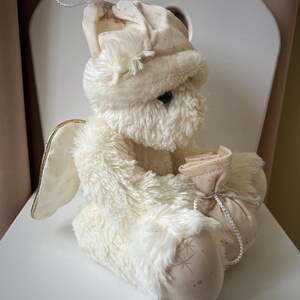May include: A plush, cream-colored angel doll with a soft, furry texture. The doll wears a matching hat and holds a small pouch. Gold-trimmed wings and star patterns on the fabric add detail. A silver string is attached to the hat.