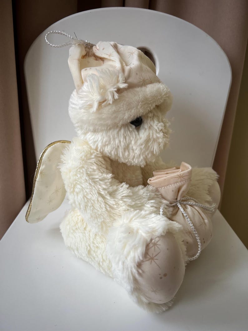 May include: A plush, cream-colored angel doll with a soft, furry texture. The doll wears a matching hat and holds a small pouch. Gold-trimmed wings and star patterns on the fabric add detail. A silver string is attached to the hat.