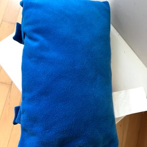 May include: A rectangular, bright blue plush pillow with a soft, fuzzy texture. The pillow features two small blue fabric tabs extending from the left side and a white, blank rectangular fabric tag attached to the right side of the item.