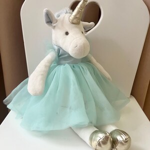 Könnte beinhalten: Eine Plüsch-Einhorn-Puppe mit weißem Körper, hellblauem Tutu-Kleid und silbernen Hufen. Das Einhorn hat ein goldenes Horn und sitzt auf einem weißen Stuhl mit herzförmigem Ausschnitt.