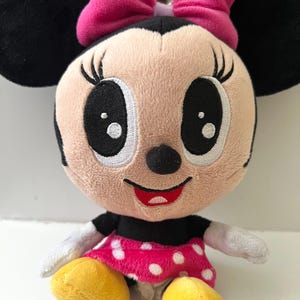 May include: A plush Minnie Mouse doll with a pink bow, black ears, and a pink polka-dot dress. The doll has large black eyes, a red smile, and yellow shoes. The soft toy is designed for children.