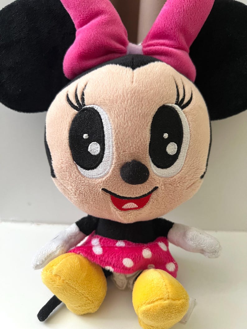 May include: A plush Minnie Mouse doll with a pink bow, black ears, and a pink polka-dot dress. The doll has large black eyes, a red smile, and yellow shoes. The soft toy is designed for children.