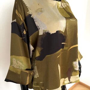 May include: Olive green blouse with a unique abstract pattern in black, beige, and white. The top features long sleeves with folded cuffs and a round neckline. The fabric has a silky sheen.