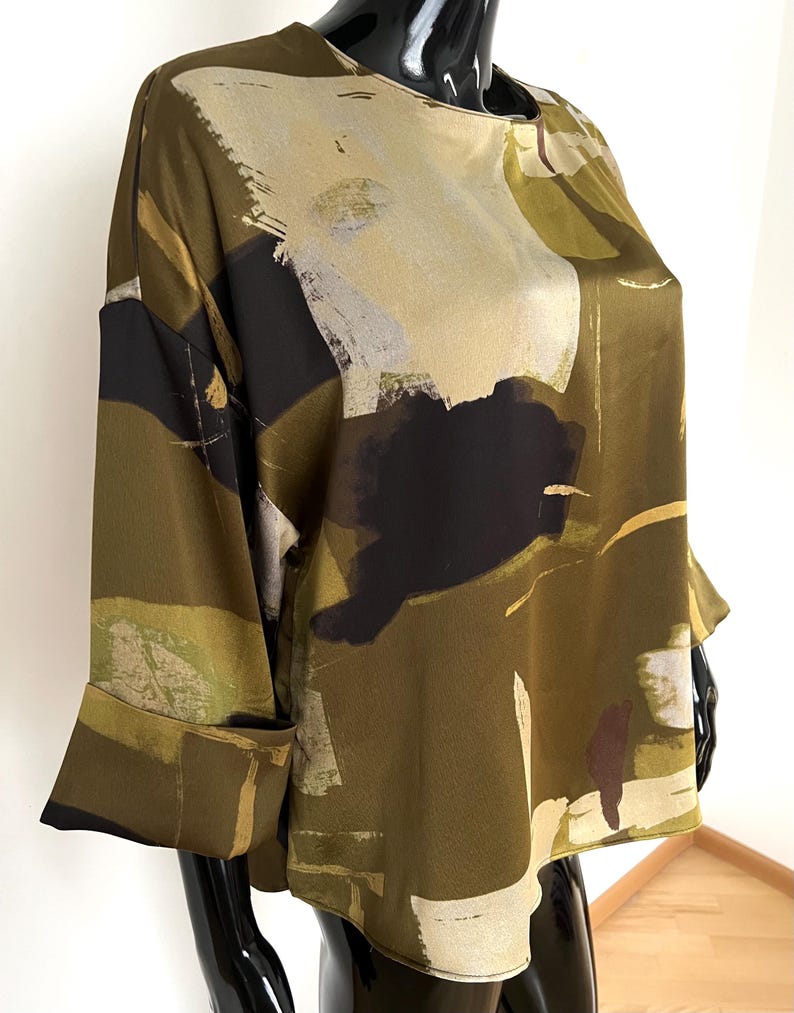 May include: Olive green blouse with a unique abstract pattern in black, beige, and white. The top features long sleeves with folded cuffs and a round neckline. The fabric has a silky sheen.