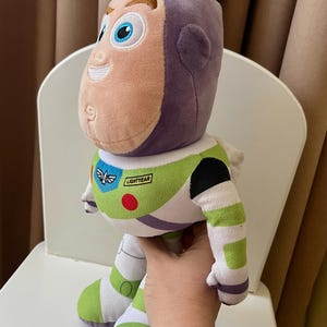 May include: A plush Buzz Lightyear doll, featuring a purple head, a light peach face, and a green and white body. The doll has a "LIGHTYEAR" patch and is sitting on a white chair.