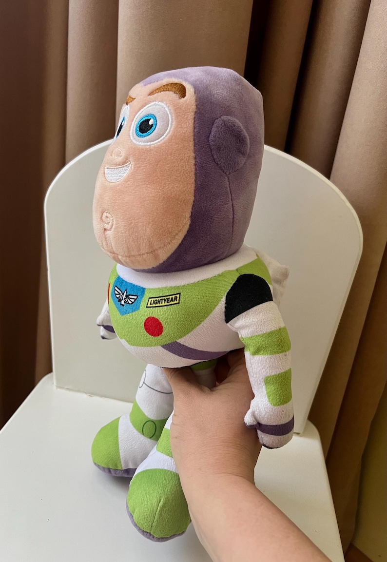 May include: A plush Buzz Lightyear doll, featuring a purple head, a light peach face, and a green and white body. The doll has a "LIGHTYEAR" patch and is sitting on a white chair.