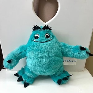 May include: A plush turquoise monster toy with black claws, hair, and accents. The toy has a smiling face with large eyes and is sitting on a white chair. The toy is made of soft, fuzzy material.