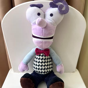 May include: A plush character toy featuring a tall, light purple head with a curled hair tuft, wide eyes, and a prominent nose. It wears a light blue long-sleeved shirt, a dark red bow tie, a black and white houndstooth vest, and dark pants.