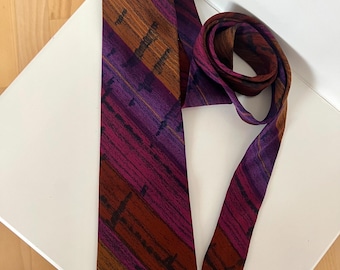 Puede incluir: Una corbata estampada en tonos morados, burdeos y marrones, con un diseño de rayas diagonales. La corbata está hecha de una tela suave y se muestra sobre una superficie blanca.