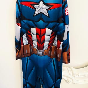 May include: A blue Captain America costume with a white star on the chest. The costume features a red and white design on the chest and a brown belt. The long-sleeved jumpsuit has the Avengers logo on the sleeve.