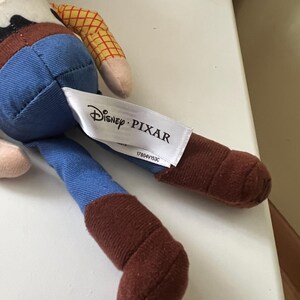 Puede incluir: Mu&ntilde;eco de Woody de Disney Pixar. El juguete presenta una camisa a cuadros amarillos, vaqueros azules y botas marrones. Una etiqueta blanca con el logo de Disney Pixar est&aacute; adjunta.