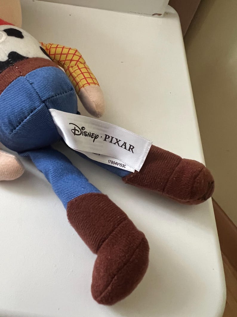 Puede incluir: Mu&ntilde;eco de Woody de Disney Pixar. El juguete presenta una camisa a cuadros amarillos, vaqueros azules y botas marrones. Una etiqueta blanca con el logo de Disney Pixar est&aacute; adjunta.