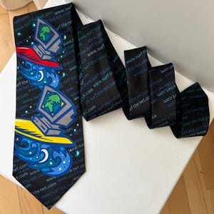 Puede incluir: Una corbata negra con un patrón repetitivo del texto www.surf the net.com en azul y blanco. La corbata muestra dos ilustraciones gráficas de un monitor de computadora que muestra un globo terráqueo, cada uno sobre una tabla de surf colorida.