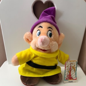Peut inclure: Poupée en peluche du personnage de Disney, Grincheux, vêtue d'une tunique jaune, d'une ceinture noire et d'un chapeau violet. La poupée a un nez et des oreilles roses et tient une petite carte avec une illustration de château et le mot "Simplelet".