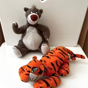 Peut inclure: Deux jouets en peluche : un ours brun avec un ventre blanc et un nez rouge, et un tigre orange avec des rayures noires. L'ours est assis et le tigre est couché. Les deux jouets sont sur une surface blanche.