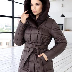 May include: A brown quilted winter coat with a fur-lined hood and a matching belt. The coat has a tailored fit and a classic design, suitable for cold weather. The coat is made of a smooth, water-resistant material.