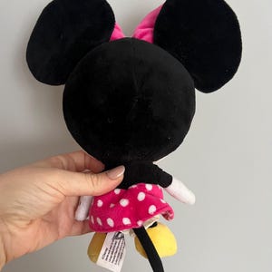 May include: A plush Minnie Mouse doll, viewed from the back, featuring large black ears, a pink bow, and a pink skirt with white polka dots. The doll has yellow shoes and a black tail. The doll is being held.