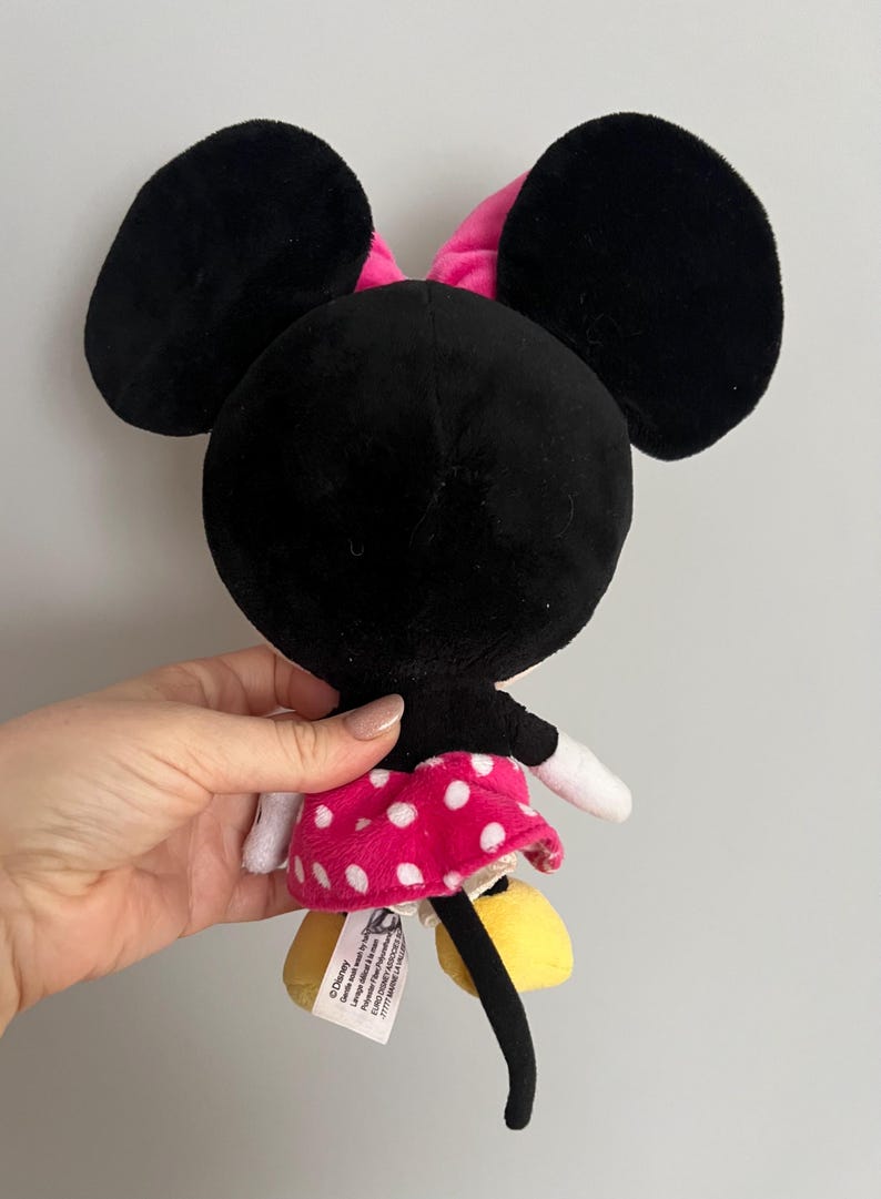 May include: A plush Minnie Mouse doll, viewed from the back, featuring large black ears, a pink bow, and a pink skirt with white polka dots. The doll has yellow shoes and a black tail. The doll is being held.