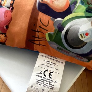 May include: A rectangular pillow featuring a colorful graphic print of Buzz Lightyear and other characters from Toy Story. The word Attic is written in black script on an orange background. A white fabric tag displays product information.