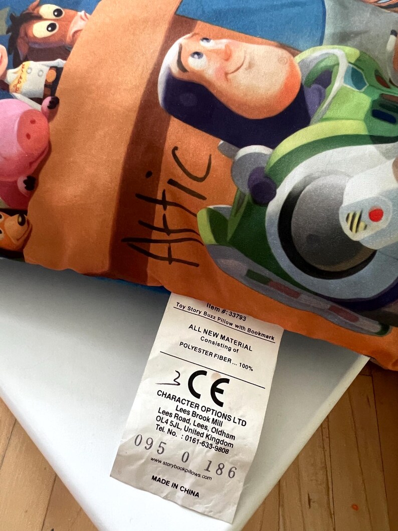 May include: A rectangular pillow featuring a colorful graphic print of Buzz Lightyear and other characters from Toy Story. The word Attic is written in black script on an orange background. A white fabric tag displays product information.