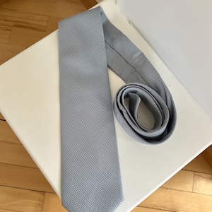 Può includere: Una cravatta azzurra con un piccolo motivo a pois ripetuto. La cravatta è appoggiata su una superficie bianca, con una parte arrotolata. La cravatta è realizzata in tessuto liscio e ha una forma classica.